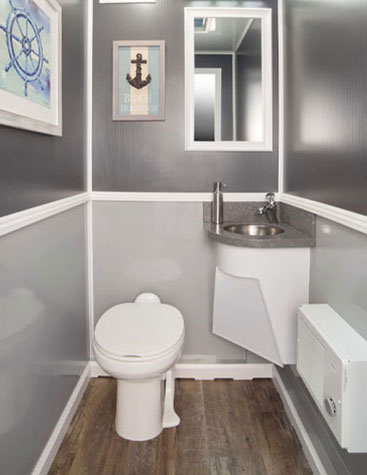 Coastal Throne Co. Casco Bay luxury restroom trailer stall interior with corner vanity, framed mirror, wood-look flooring, and nautical artwork