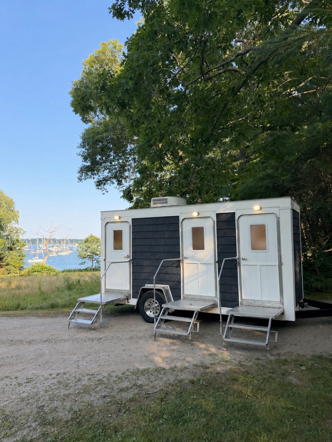 Coastal Throne Co. Pemaquid 3-stall luxury restroom trailer with navy shingled exterior parked under a shade tree overlooking a Maine harbor