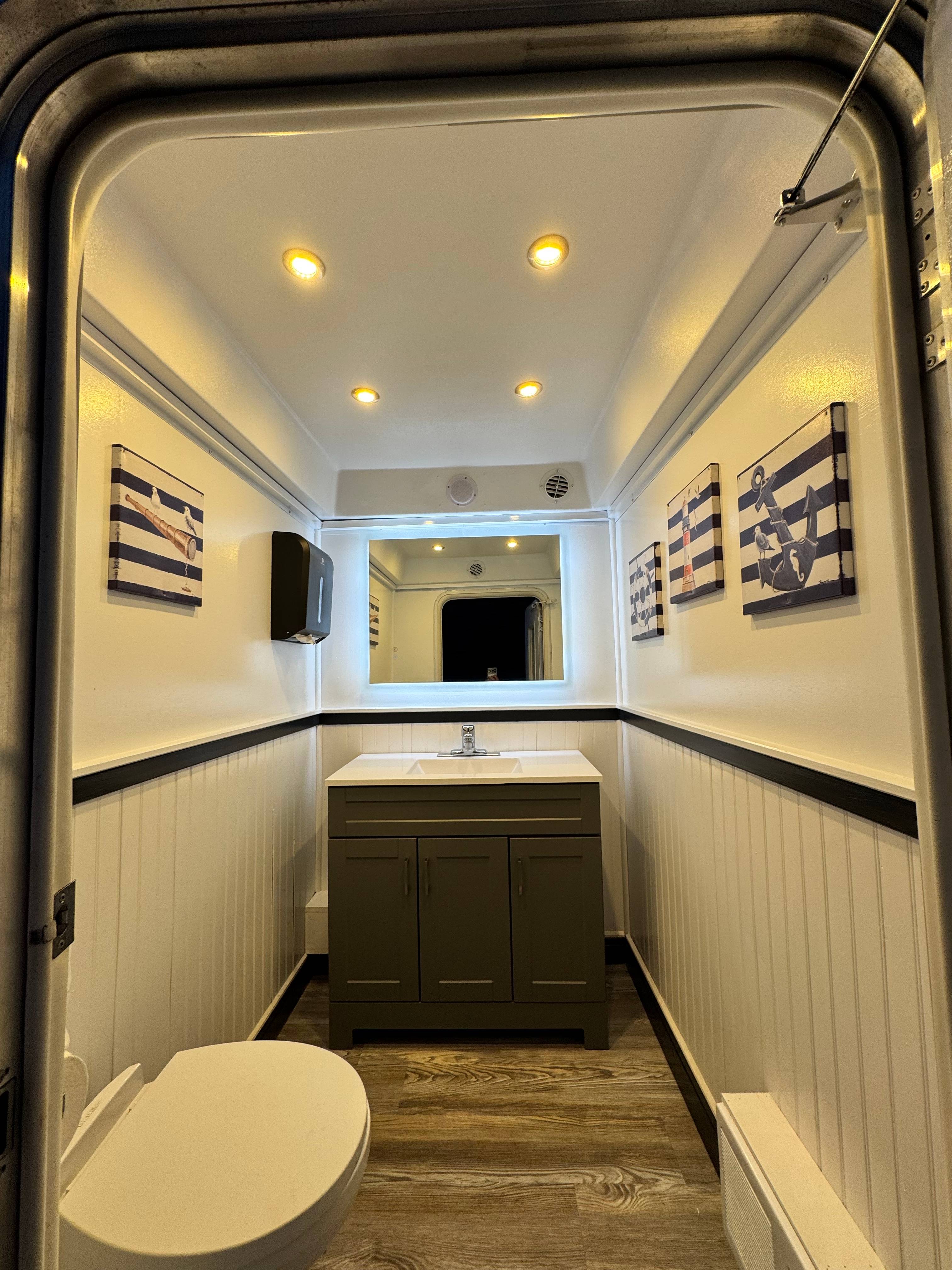 Coastal Throne Co. Pemaquid luxury restroom trailer interior with charcoal vanity, backlit mirror, white beadboard walls, wood-look flooring, and nautical framed artwork